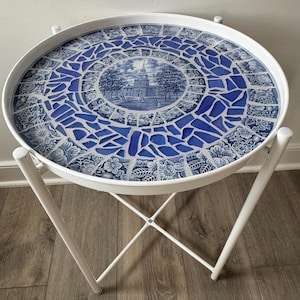 May include: White round side table with a blue and white mosaic top. The mosaic design features a central image of a building with trees and a spire. The table has a white metal frame with four legs.