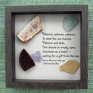 Può includere: Un'opera d'arte incorniciata con vetro marino e una citazione. La cornice grigia circonda uno sfondo bianco sporco. I pezzi di vetro marino sono di vari colori, tra cui beige, viola e verde. La citazione recita: "Pazienza, pazienza, pazienza...aspettando un dono dal mare."