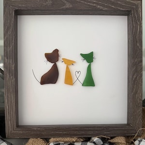 Puede incluir: Obra de arte enmarcada con tres gatos estilizados en marrón, amarillo y verde, unidos por una cola en forma de corazón. La obra está sobre un fondo blanco dentro de un marco de madera marrón oscuro.