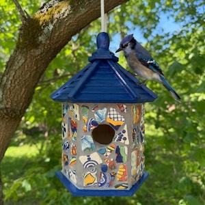 Pode incluir: Uma casa de pássaros artesanal com telhado azul e paredes de mosaico de azulejos coloridos, pendurada num galho. Um gaio-azul está pousado no telhado. A casa de pássaros é um adorno decorativo para o jardim.