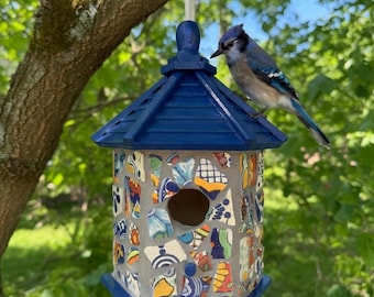 May include: A handcrafted birdhouse with a blue roof and mosaic tile walls, hanging from a tree branch. The mosaic features colorful ceramic pieces. A blue jay perches on the roof, adding a touch of nature to the decorative birdhouse.