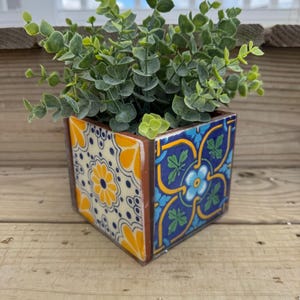 Puede incluir: Un macetero cuadrado de cerámica con un diseño de azulejos geométricos coloridos en tonos azules, amarillos y blancos. El macetero está lleno de una planta verde de hojas.