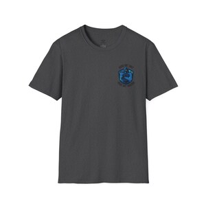 May include: A dark heather grey t-shirt with a graphic of a hooded figure in a blue and green shield. The text "GOOD ART SHIT GOOD ART FRIENDS" is printed around the shield.