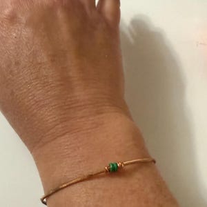 Könnte beinhalten: Ein kupferfarbenes Drahtarmband mit einer kleinen grünen Perle und einer kupferfarbenen Perle. Das Armband wird am Handgelenk getragen. Schlichtes, minimalistisches Design.