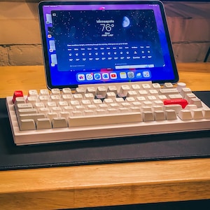 May include: A white wireless keyboard with red keycaps on a wooden desk. A tablet with a weather app showing the temperature in Minneapolis, Minnesota as 76 degrees Fahrenheit is in the background.