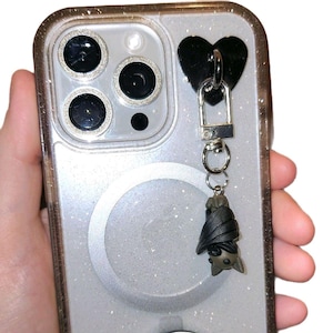 May include: A clear glitter phone case with a black heart-shaped charm and a silver clasp. A small, dark gray bat charm hangs from the clasp. The phone case has a silver magnetic ring on the back.
