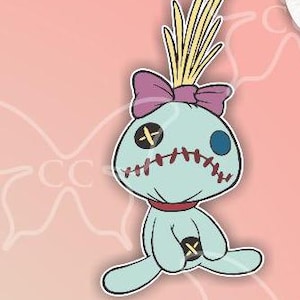 May include: A light blue cartoon doll sticker with a purple bow, yellow hair, and red stitching. The sticker is 5" x 9" in size. The doll has a black cross for an eye and a blue eye on the other side.