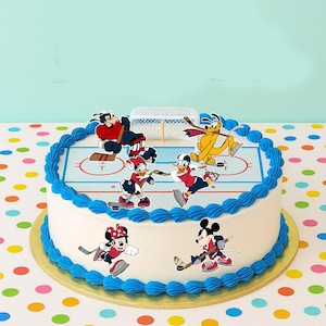 Puede incluir: Un pastel redondo blanco con borde de glaseado azul y una base dorada. El pastel está decorado con personajes de dibujos animados jugando al hockey sobre una pista de hielo azul y blanca. Los personajes incluyen Mickey y Minnie Mouse, Donald Duck y Pluto.