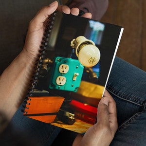May include: A spiral bound notebook with a photo of an electrical outlet and a light bulb. The outlet is teal blue and the light bulb is white. The photo is on a wooden surface with other objects.