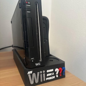 Könnte beinhalten: Schwarze Wii-Konsole mit passendem Ständer. Der Ständer trägt den Schriftzug "Are Wii Gonna Have A Problem" mit roten und blauen Fragezeichen. Die Konsole steht auf einer Holzoberfläche.