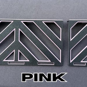 May include: Two dark gray metal pieces with a geometric design and pink accents. The larger piece has a peace sign shape. The word "PINK" is displayed in bold, white letters at the bottom.