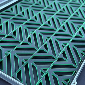 May include: A geometric design in a rectangular frame. The design features a repeating pattern of black shapes with green accents. The frame is silver. The overall aesthetic is modern and abstract.