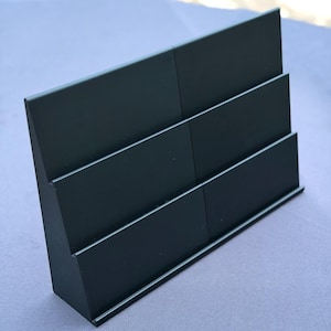 May include: A dark green, three-tiered display stand. The stand is made of a solid material and has a rectangular shape. The tiers are separated by horizontal lines, creating individual compartments. The stand is designed to hold and display items.
