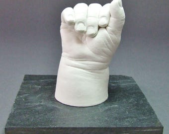 May include: A white plaster cast of a baby's clenched fist, mounted on a dark gray rectangular base. The hand is detailed, showing the knuckles and the curve of the fingers. The base is approximately 6 inches long.