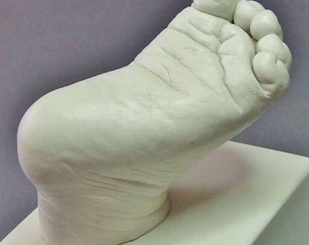 May include: A white plaster cast of a baby's foot, showcasing detailed wrinkles and the shape of the toes. The foot is positioned on a white rectangular base, with a soft, smooth texture. The overall impression is of a delicate keepsake.