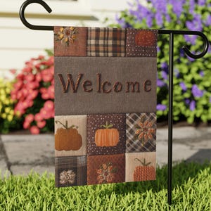 May include: A rectangular garden flag hanging on a black metal stand. The flag features a patchwork design with brown, orange, and black squares displaying pumpkins and floral patterns. The word Welcome is centred in brown text on a grey band.