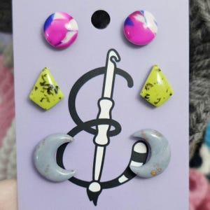 May include: A collection of handmade earrings in various shapes and colors. Includes pink and white marbled round studs, yellow diamond-shaped earrings with green speckles, and gray crescent moon earrings. The earrings are displayed on a purple card with a crochet hook graphic.