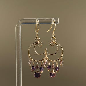 Könnte beinhalten: Goldfarbene Creolen mit hängenden lila Perlen. Die Ohrringe haben ein Doppelreifen-Design mit kleinen, zarten lila Perlen und größeren, runden Perlen, die von unten herabhängen. Die Ohrringe werden auf einem transparenten Ständer vor neutralem Hintergrund präsentiert.