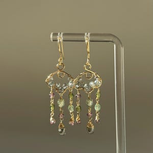 May include: A pair of gold-toned wire earrings with a cloud-like design. The earrings feature dangling strands of light blue, green, and pink beads, with a teardrop-shaped bead at the end of each strand. The earrings are displayed on a clear stand.