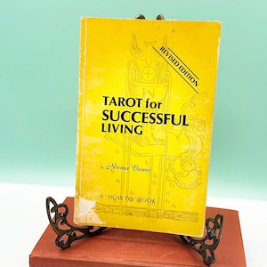 May include: A yellow book titled "TAROT for SUCCESSFUL LIVING" with "REVISED EDITION" text, displayed on a decorative bronze stand atop a red book. The book is a "HOW TO" book by Norma Cowie.