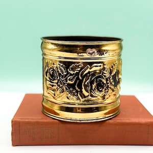 Peut inclure: Un récipient en métal doré avec un motif floral de roses détaillé. Le récipient est cylindrique et repose sur un livre marron. Le fond est de couleur vert clair.