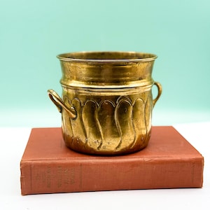 May include: A vintage brass planter with a wavy pattern and small handles. The planter sits atop a closed, red-orange book. The background is a solid, light teal color.