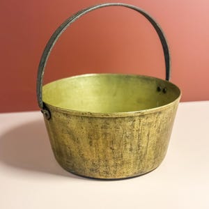 Puede incluir: Un cubo de latón vintage con un asa de metal curvada. El cubo tiene forma redonda y una superficie ligeramente texturizada. El interior del cubo es de un tono más claro de latón. El fondo es de color marrón rojizo.