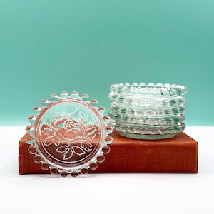Puede incluir: Dos objetos decorativos de cristal transparente. Uno es un posavasos redondo con un diseño de rosa y un borde de cuentas. El otro es un pequeño recipiente redondo con cuentas. Ambos están sobre un libro vintage con tapa roja.