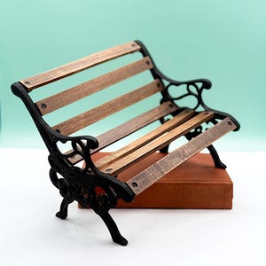 Op de afbeelding: Miniatuur bankje met een zwart gietijzeren frame en houten latten. Het bankje staat op een bruin boek, tegen een lichtblauwe achtergrond. Een decoratief item.