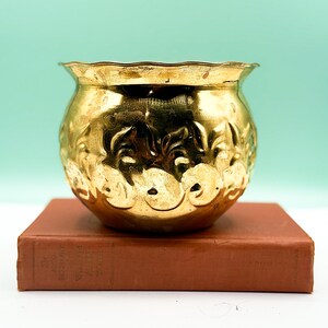 Pode incluir: Uma pequena tigela redonda de metal dourado com uma borda ondulada. A tigela tem uma superfície texturizada com um padrão floral repetido. Está em cima de um livro castanho com texto desbotado na lombada. O fundo é de cor verde-azulado claro.