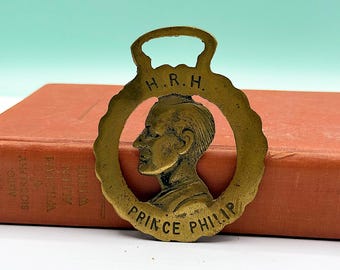May include: A circular brass horse brass featuring a profile relief of Prince Philip. The text H.R.H. is engraved at the top and PRINCE PHILIP is engraved at the bottom. The brass has a loop at the top for attachment.