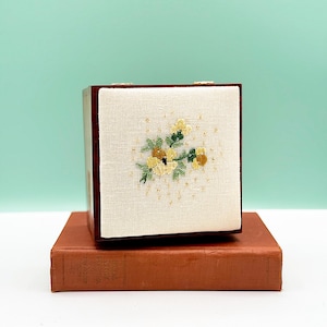 May include: A small, square box with a cream-colored fabric front featuring a floral embroidery design in yellow, green, and brown. The box is set on top of a vintage, reddish-brown book. The background is a light teal color.