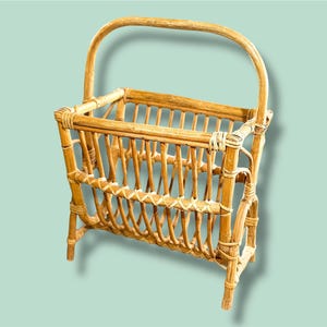 May include: A vintage rattan magazine rack with a curved handle. The rack is made of woven light brown rattan with a rectangular shape and vertical slats. The handle is arched, and the piece has a natural, rustic aesthetic.
