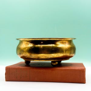 Może przedstawiać: Mała, zabytkowa mosiężna miska o zaokrąglonym kształcie i trzech małych nóżkach. Miska stoi na zabytkowej, czerwonobrązowej książce. Tło jest w miękkim, jasnym, turkusowym kolorze.