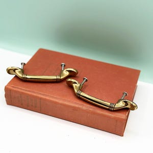 Op de afbeelding: Twee gouden kastdeurgrepen met een decoratief ontwerp. De grepen liggen op een bruin boek.