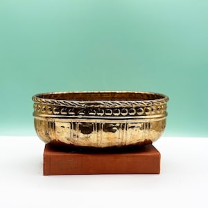 Puede incluir: Un cuenco dorado de forma ovalada con una superficie texturizada y un borde en forma de cuerda. El cuenco está sobre un libro marrón sobre un fondo verde claro. El cuenco tiene una estética vintage.