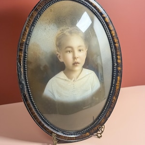 Puede incluir: Retrato ovalado antiguo en un marco de madera oscura con un borde decorativo. El retrato presenta una imagen en tonos sepia de un niño con una camisa blanca. El marco está sostenido por un soporte de metal decorativo.