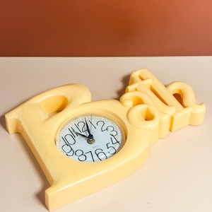May include: A cream-coloured decorative clock in the shape of the word "Paris". The clock face is white with black numerals and hands. The clock is made of a solid, smooth material and sits on a light-coloured surface.