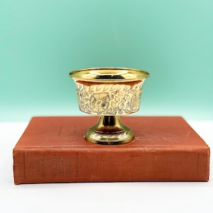 May include: A small, ornate brass cup with a floral design, set on a vintage book. The cup features a wide rim and a pedestal base. The book is reddish-brown, against a light teal backdrop.