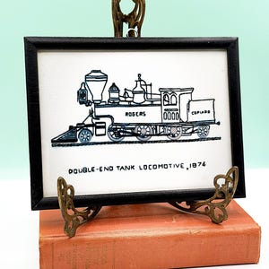 Può includere: Opera d'arte incorniciata in nero di una locomotiva a serbatoio a doppia estremità, datata 1874. L'illustrazione in bianco e nero è esposta su sfondo bianco. L'opera è appoggiata su un supporto decorativo in ottone, appoggiato su un libro d'epoca.