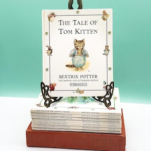 Peut inclure: Une pile de livres pour enfants, dont "The Tale of Tom Kitten" de Beatrix Potter, exposée sur un présentoir décoratif en métal. Les livres présentent des illustrations et du texte colorés. Les livres sont empilés sur un livre rouge.