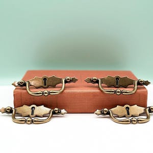 Pode incluir: Quatro puxadores de gaveta de latão antigo com um design de buraco de fechadura. Os puxadores estão dispostos sobre uma capa de livro vermelha.