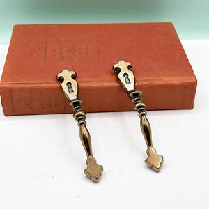 Puede incluir: Dos tiradores de armario de latón antiguo con un diseño de ojo de cerradura. Los tiradores tienen un diseño decorativo y ornamentado y están en buen estado.