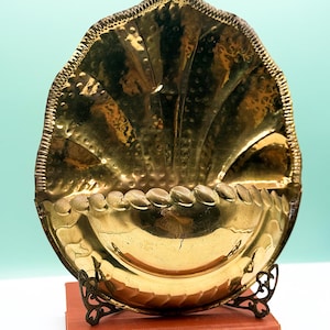 May include: A vintage brass ashtray, shaped like a shell, with a textured surface and decorative edging. It's supported by a small, ornate metal stand, resting on a red book. The design combines practicality with artistic flair, a classic piece of decor.