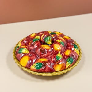 May include: A round ceramic pie dish with a decorative lid featuring a raised, hand-painted design of multiple red and yellow apples with green leaves. The dish has a scalloped, ruffled edge in a muted yellow tone, creating a textured appearance.