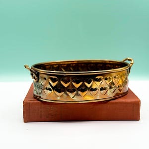 May include: An oval, gold-coloured metal container with heart-shaped cutouts. It features two small handles and rests on a vintage, reddish-brown book. The background is a solid, light teal colour.