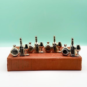 Puede incluir: Una colección de botellas de vino, copas y decantadores dorados en miniatura se colocan sobre un libro antiguo. Los artículos se exhiben sobre un fondo verde azulado claro, creando una disposición visualmente atractiva. La portada del libro es de color marrón rojizo cálido.