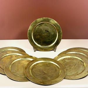 May include: A set of six circular brass plates with a polished, reflective finish and a wide, flat rim. One plate is positioned upright on a small metal stand, while the other five plates are arranged in a semi-circle in front of it.