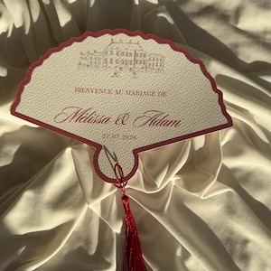 Peut inclure: Un faire-part de mariage en forme d'éventail de couleur crème, avec une bordure rouge festonnée et un pompon rouge. L'invitation comprend le texte "Bienvenue au Mariage de Melissa & Adam" et la date "27.07.2026".