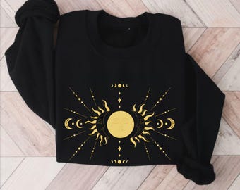 May include: Black sweatshirt with a gold sun and moon design. The design features a sun and moon with faces, surrounded by crescent moons and stars.